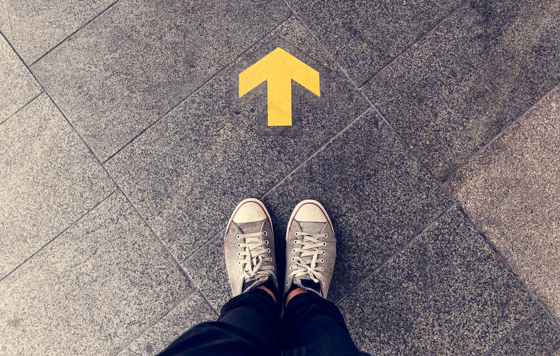 Symbolising Website Breadcrumbsthere is a person wearing white trainers, stading before a yellow direction arrow that is on the ground. 