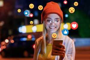Business Growth Through Facebook - this image shows a lady at night with an ornage hat and glasses, she is looking at her mobile phone and smiling at there are facebook icons around the phone