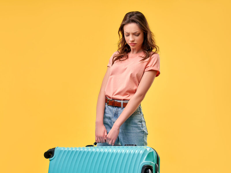 Why Your Website Is Carrying Extra Baggage - this image shows a bright yellow background with a lady in the foreground with ba very heavy suitcase, concept for website carrying excess baggage