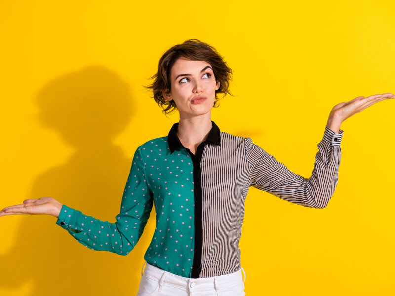 The Difference Between Content That Educates and Content That Converts this image shows a bright yelow background with a lady in a colourful blouse in the foregroud looking thoughtful, she has her arms out to the sides and raised one higher than the other as if balancing or measuring options.